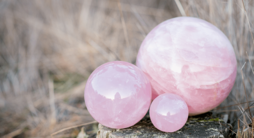 Three polished rose quartz spheres of varying sizes placed on a rock with a blurred natural background of dry grass.