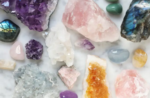 A diverse collection of crystals and gemstones, including amethyst, rose quartz, citrine, and labradorite, displayed on a marble surface.