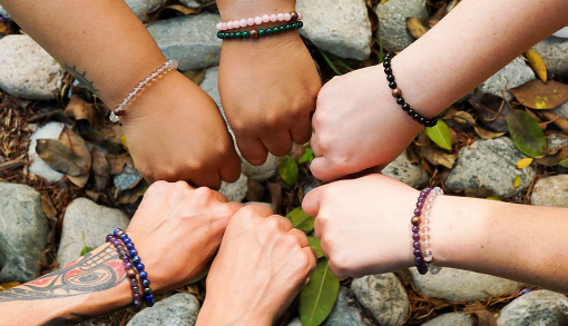 Six hands wearing different healing crystal bracelets, forming a circle over a ground covered with rocks and leaves